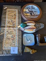 Top view of lot featuring framed embroidery, decorative plates, carved soapstone bird, porcelain duck, cast iron owl figurine, and two small brass bird figurines.