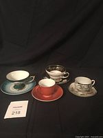 Four china cups and saucers shown grouped on dark background, varied colors and patterns.