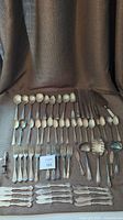 Array of silver plated cutlery including spoons, forks, knives, and serving utensils laid out on a brown fabric background.