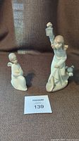 Photo showing both Lladro Nao porcelain figurines, the kneeling angel on the left and the taller girl with lantern and dog on the right on a brown fabric surface.