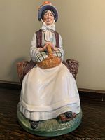 Full view of the Royal Doulton porcelain figurine showing seated elderly woman holding a basket on a bench with a green base and small bird detail.
