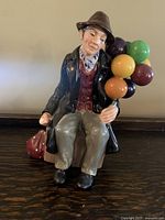 Front view of Royal Doulton bone china figurine showing man seated holding colorful balloons with a small bag beside him.