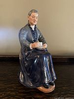 Front view of the bone china figurine showing the elderly woman sitting holding a teacup and saucer, dressed in dark blue with gray jacket, detailed face and hair.