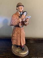Front view of the Royal Doulton "The Detective" figurine showing the detective holding magnifying glass and paper, wearing brown quilted coat and deerstalker hat.