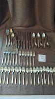 Full set of cutlery laid out on brown fabric showing all pieces: knives, forks, spoons and serving utensils.