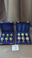 Two open blue velvet-lined cases each containing six tarnished silver plated tumblers with gold-tone clasps on cases