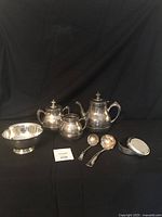 Full view of all silver plated items including tea set, bowl, ladles, and trinket box arranged on black backdrop.