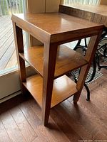 Front and side angle of small wooden table showing top and two shelves below, placed by a glass door.