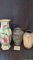 Three vases displayed on a black background: left - tall hexagonal Asian motif ceramic vase, center - clear ribbed glass vase, right - shimmering multi-colored ceramic vase.