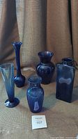 Collection of 5 blue glass vases pictured together on a brown fabric backdrop.