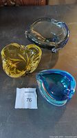 Three vintage hand-blown glass ashtrays displayed together including amber heart-shaped, blue/grey round, and blue teardrop-shaped ashtrays.