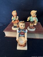 Front view of three Hummel figurines on a book: Girl with bowl kneeling in front, Boy with horse holding a mule figurine, and Girl with doll figurine in back.