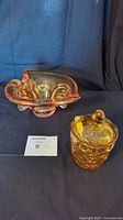 Shot showing both amber glass ashtray and sugar holder with lid and spoon against a dark cloth backdrop