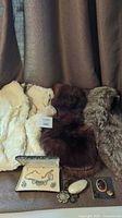Three genuine fur items: off-white fur vest, brown/black fur stole collar, and white/grey fur stole collar shown together with assorted vintage jewelry items positioned in front including brooches, necklaces, and pins.