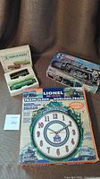 Lot displayed on brown fabric including Lionel train clock in original box, vintage tramway wagons box with two miniature tram cars visible, and Hudson locomotive model kit box.