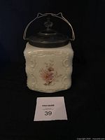 Front view of vintage biscuit jar showing embossed floral design, painted flowers, and silver plated lid with handle