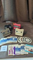 Four Viewmaster viewers (two beige, one black, one red) with multiple reels and packaging laid out on fabric surface.