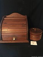 Full view of the vintage wooden roll top breadbox with roll-top cover and white knob, alongside the vintage teak lidded wooden vessel, both placed against a black background.
