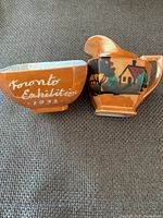 Front view of a cream and sugar set with orange glaze and white text 'Toronto Exhibition 1932' beside a matching orange decorative item with a house silhouette design.
