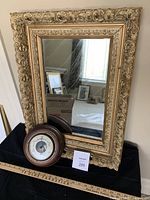 Full view of the vintage gilded mirror standing upright with Taylor barometer placed in front on a black surface.