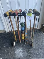Full view of six wooden croquet mallets with striped heads and handles, standing upright in a black metal stand on wheels. Visible wood grain and wear, faded paint, and rubbed ends on mallets.