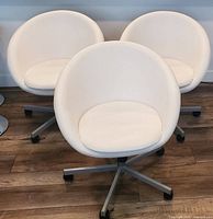 Three white faux leather rounded office chairs with curved backs and separate seat cushions. Chairs stand on metal bases with caster wheels on a wood floor.