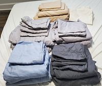 Five folded sets of queen size bed sheets and two pillow covers in various colors including beige, light gray, white, light blue, and dark gray, arranged on a bed.