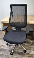 Front view of ergonomic black office chair showing mesh backrest, cushioned black seat, adjustable armrests, and rolling casters.