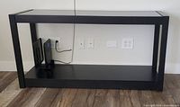 Front view of a black TV console with glass top on a wooden floor against a wall, showing the open lower shelf, power outlets behind and no WiFi unit present.