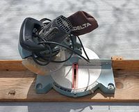 Close-up photo of Delta compound miter saw showing circular blade, handle, and brown dust collection bag.
