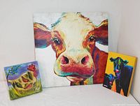 Lot of three colorful farmyard cow canvas art pieces against a white background.
