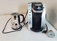 Nespresso frother and Keurig coffee maker with cords and removable filter shown on white surface.