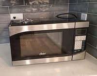 Front angled view of Oster stainless steel and black countertop microwave on kitchen counter, showing the door, keypad, and display.