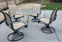 Patio set with round glass top table and 4 swivel rocker chairs arranged on concrete patio outdoors.