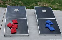 Two opened cornhole game boards on concrete surface with four red and four blue beanbags placed on top.