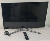 Front view of the Samsung flat panel TV with stand and remote control placed on table. TV is off.