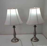 Pair of silver tone table lamps, each with white fabric Stiffel shade, shown on white surface against white wall.