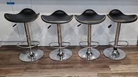 Front view of 4 chrome bar stools with black seats lined up side by side on wood floor.