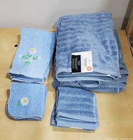 Set of 2 new blue bath towels and 2 new blue washcloths stacked, with 2 vintage blue hand towels and 2 vintage blue washcloths featuring white daisy embroidery laid next to them on a wooden surface.