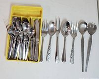 Overall view of flatware including forks, spoons, knives in yellow plastic flatware organizer tray, plus additional serving forks, spoons, and salad utensils lined out beside tray.