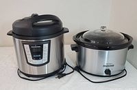Fairwind stainless steel electric pressure cooker on the left and Rival stainless steel crock pot slow cooker on the right, both plugged in and ready to use