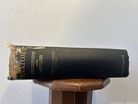 Side view of book showing black cloth hardcover with gold text on spine and tape repair at top.