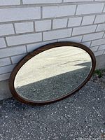 Antique oval mirror leaning against light brick wall on a concrete surface. Visible wooden frame and mirror condition clear.
