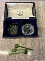 Top view of the opened box showing two crystal glass bowls, one green and one clear, placed inside separated by a divider with space for spoons in the corner. Three small green glass spoons displayed outside case.