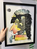 Full view of the framed surrealist mixed media painting on white wall, held by hand showing scale