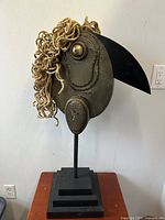 Full frontal photo of large grotesque bird sculpture with prominent black beak and rope hair detail, mounted on tiered black wooden base.