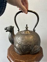 Silver-plated teapot shaped like a camel, showing the full length view with handle upright and detailed body embossing.
