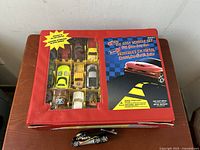 Red fitted carrying case with 9 Hot Wheels toy cars visible through window, containing various car models including yellow and white cars and a black car with flames outside the case.
