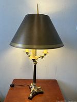 Front view of Victorian tri-footed table lamp with dark metal conical shade, showing ornate brass base and three faux candle bulbs lit.