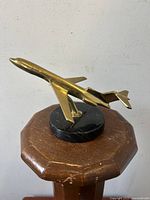 Side view of brass jet airplane figurine on wooden stand showing polished brass surface and black marble base.